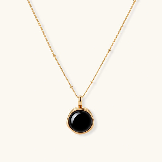Circular black agate pendant on gold-plated sterling silver chain against cream background, showing modern minimalist design.